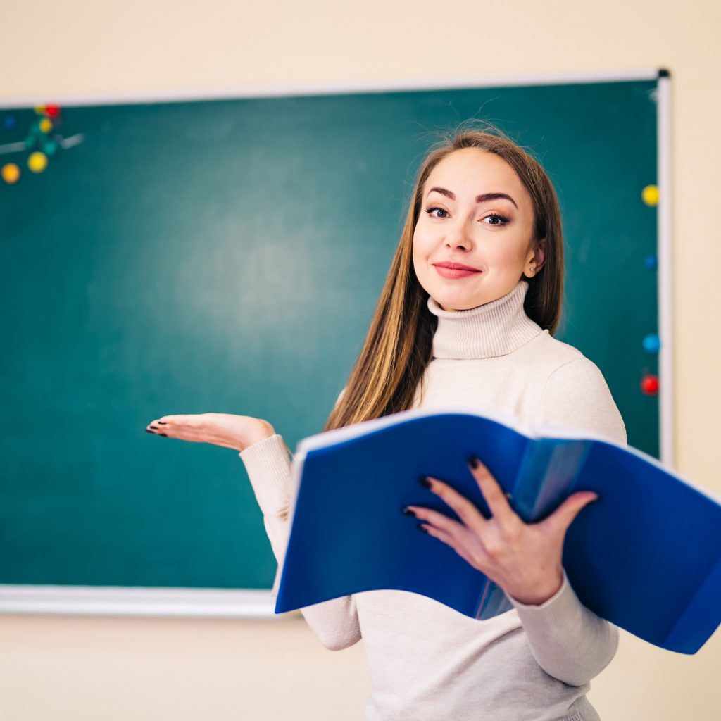 young-teacher-is-standing-near-blackboard-classroom-showing-i-don-t-know-with-hands-she-reading-notes-classroom (1) (2)